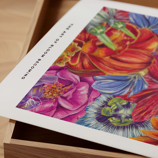 Close up of colorful floral artwork paper print with text that reads The Art Of Bloom Becoming on a wooden surface