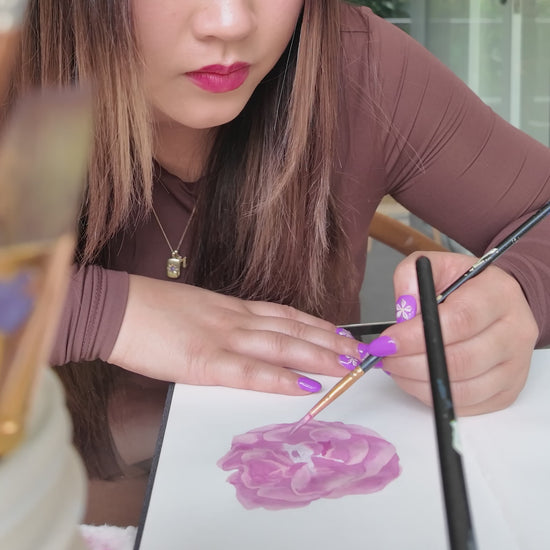 Girl in brown top holding a paint brush painting a pink rose flower