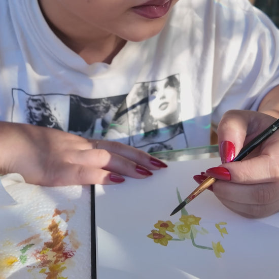Girl in white top holding a paint brush painting a yellow flower