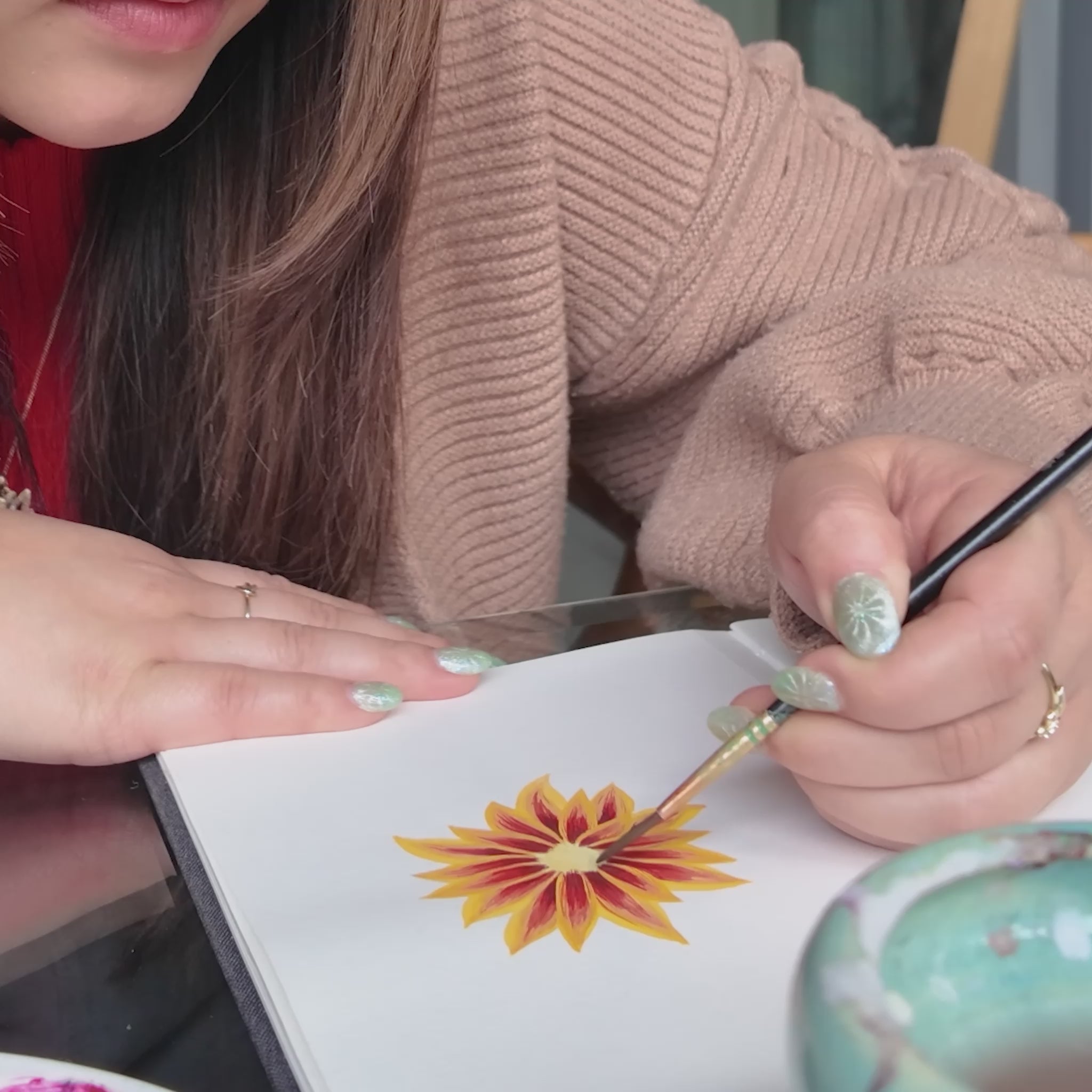 Girl in red sweater holding a paint brush painting a red and yellow flower